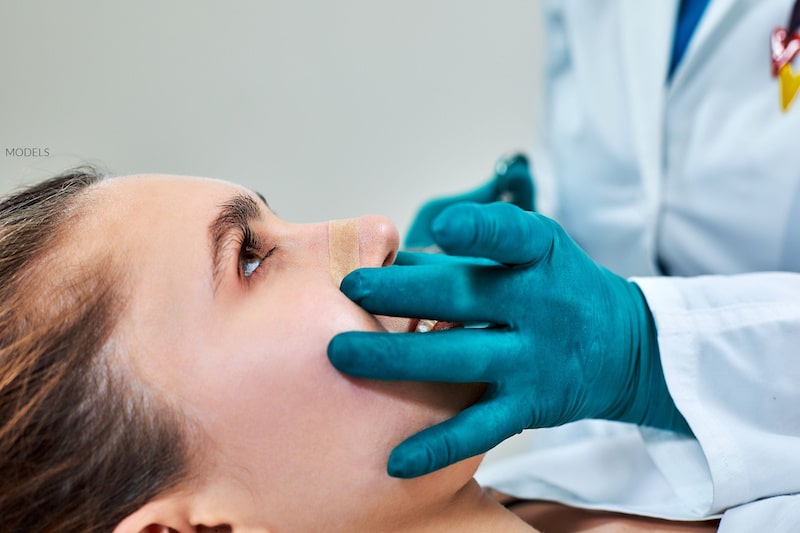 A female patient smiling while a plastic surgeon examines her nose