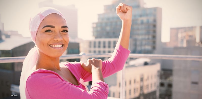 Happy woman wearing pink and a head scarf showing her strength with the impression that she is a breast cancer survivor.